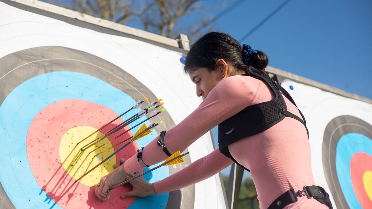 A female archer adjusts arrows on a target during a sunny day, showcasing precise archery skills.